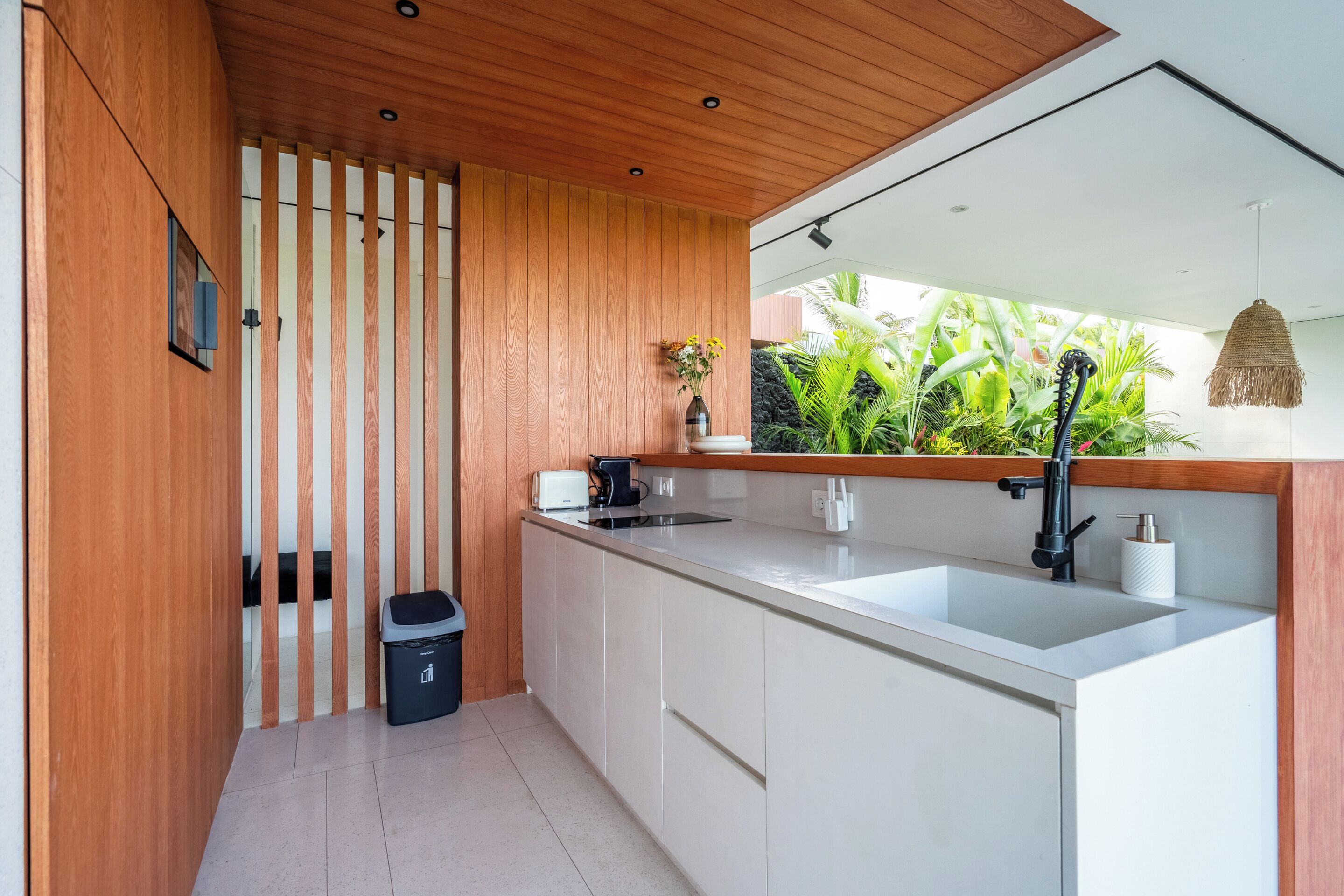 A modern kitchen with white cabinets, a wooden ceiling, and a view of lush greenery outside.