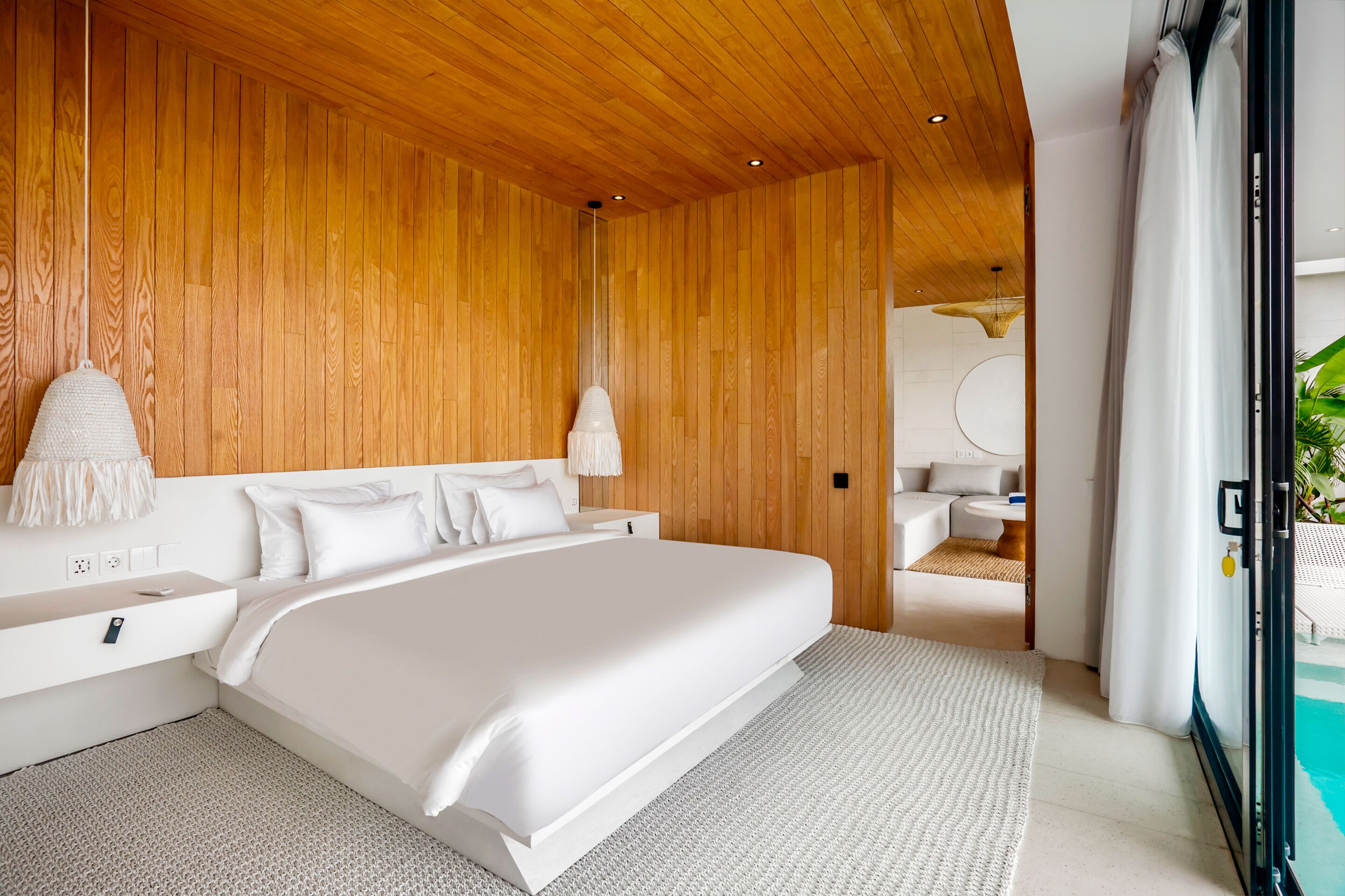 A modern bedroom with a wooden ceiling, a large bed, a bedside table, and a view of a pool area.