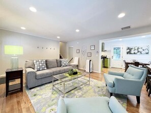 A living room with a sofa, armchairs, a coffee table, a lamp, and a kitchen visible through an open door.