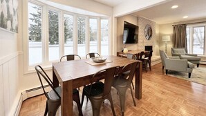 Amplia sala de estar con chimenea, comedor con mesa y sillas de madera, y ventanas grandes con vista a árboles.