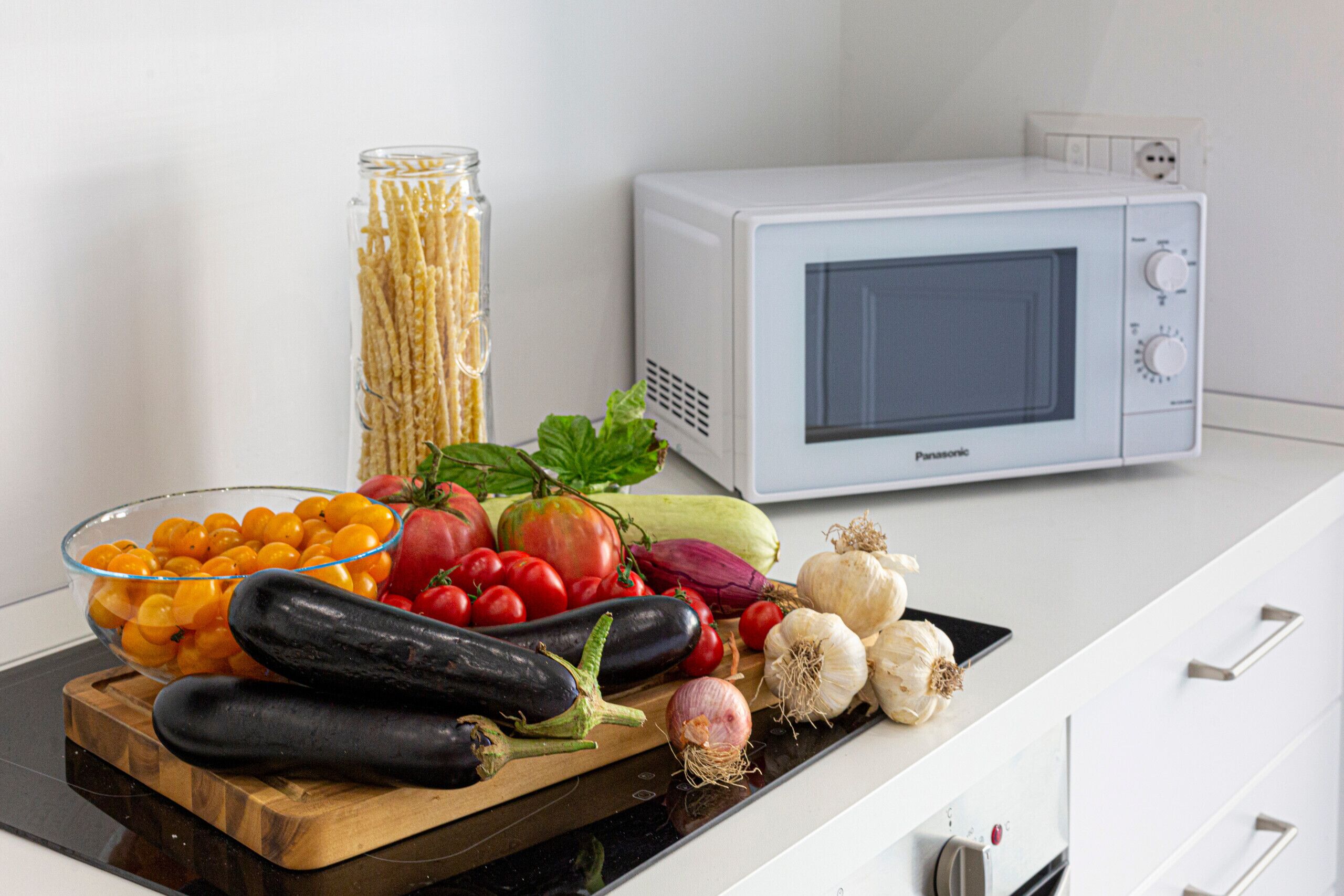 Uma bancada de cozinha com diversos vegetais frescos, uma tábua de corte e um micro-ondas branco.