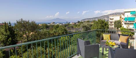 Un balcón con mobiliario de mimbre, una mesa con frutas y vistas a la montaña y al paisaje urbano.