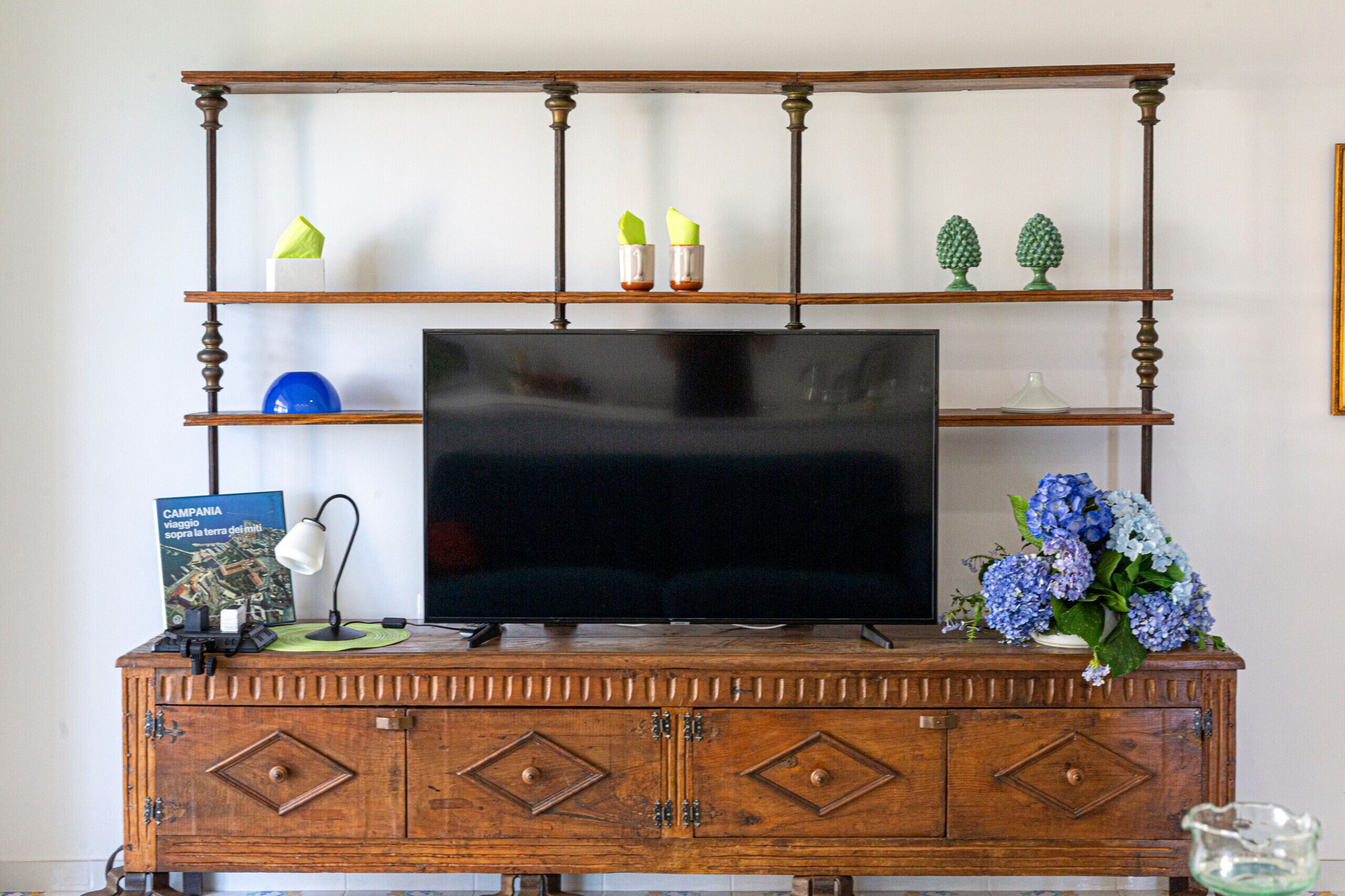 A wooden entertainment center with a flat-screen TV, a book titled 'Campania', a lamp, and a vase with flowers.