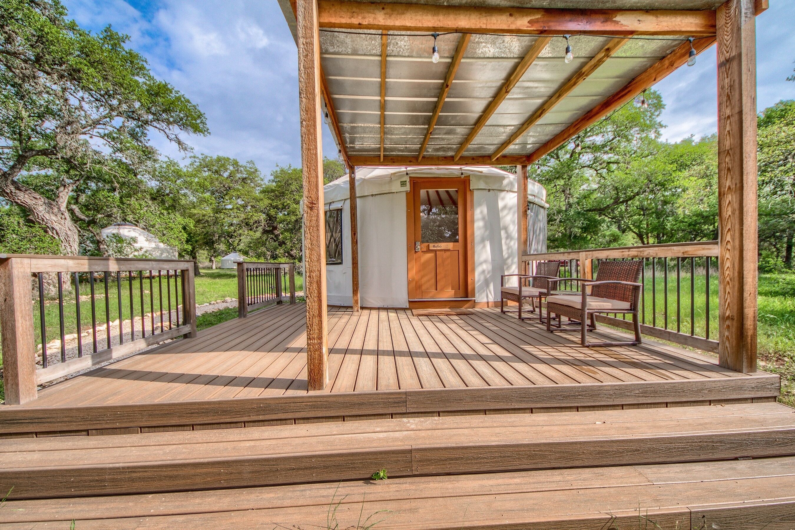 A wooden deck with a covered area, a white building with a door, and outdoor furniture.