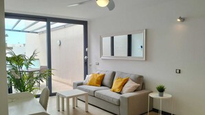 A modern living room with a grey sofa, white coffee table, and a small side table with a plant. There is a ceiling fan, a large window, and a view of the outside. - Beachfront apartment, with sea and pool view, in El Cotillo, Fuerteventura (El Cotillo, La Oliva)
