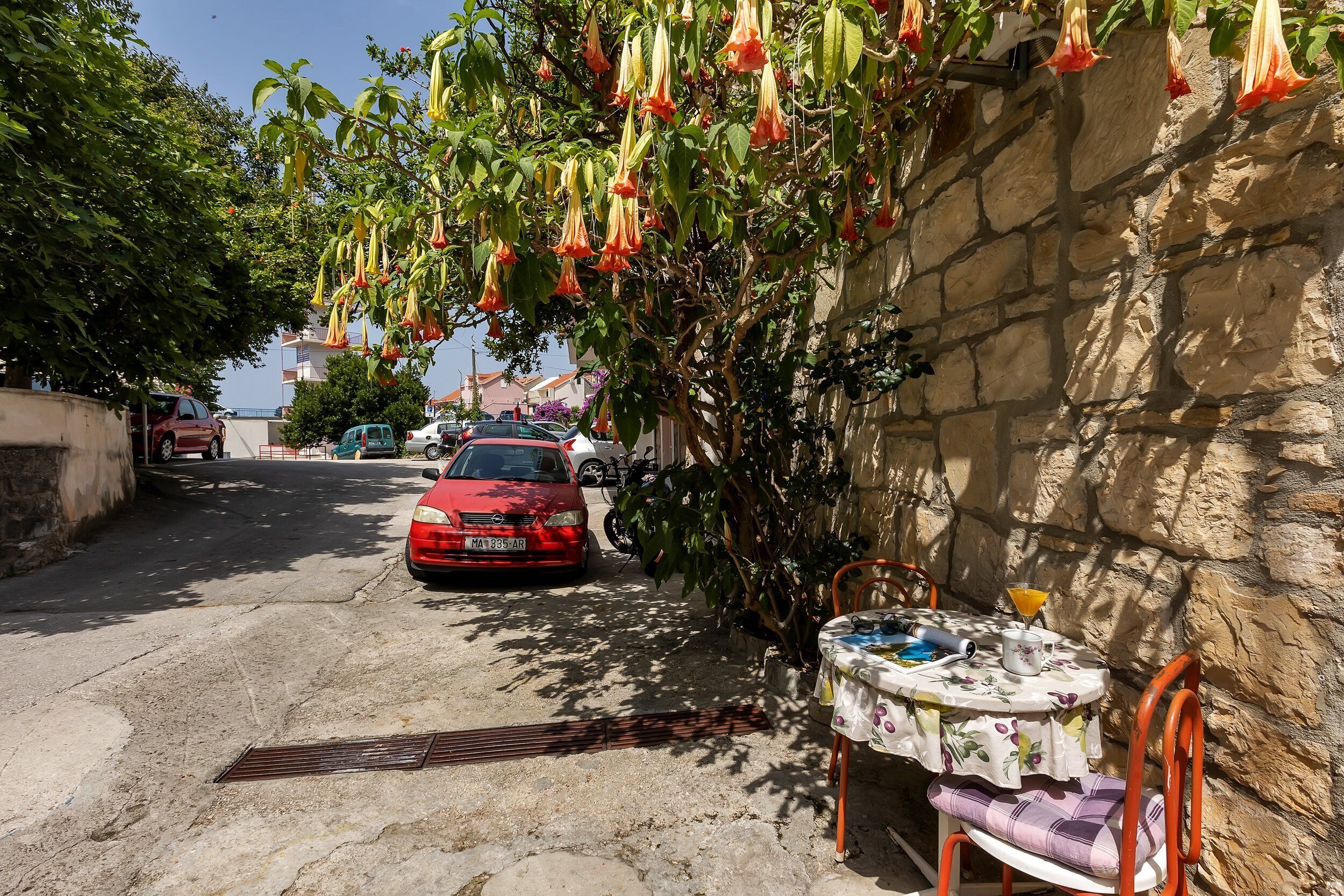 A narrow street with parked cars, a table with chairs, and hanging orange flowers.