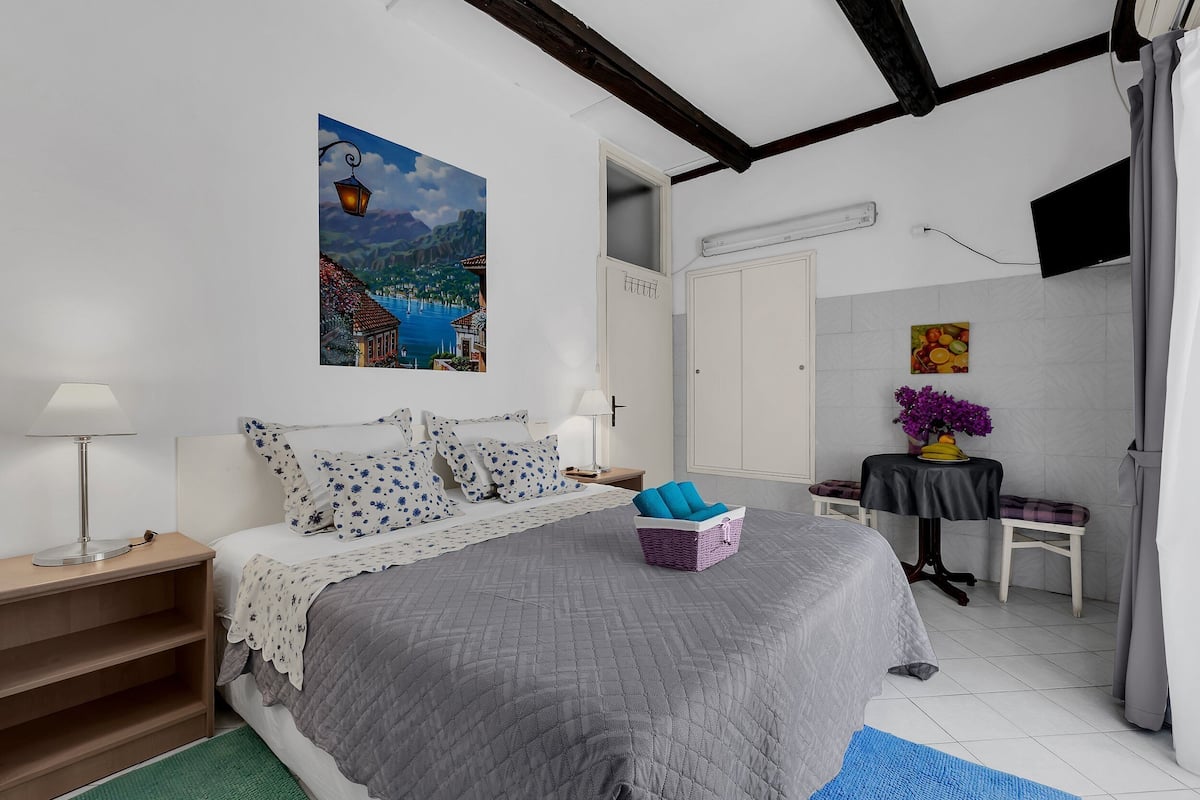 Une chambre à coucher avec un lit, une table de chevet, une lampe, un tableau, une table ornée d’un vase de fleurs et une chaise.