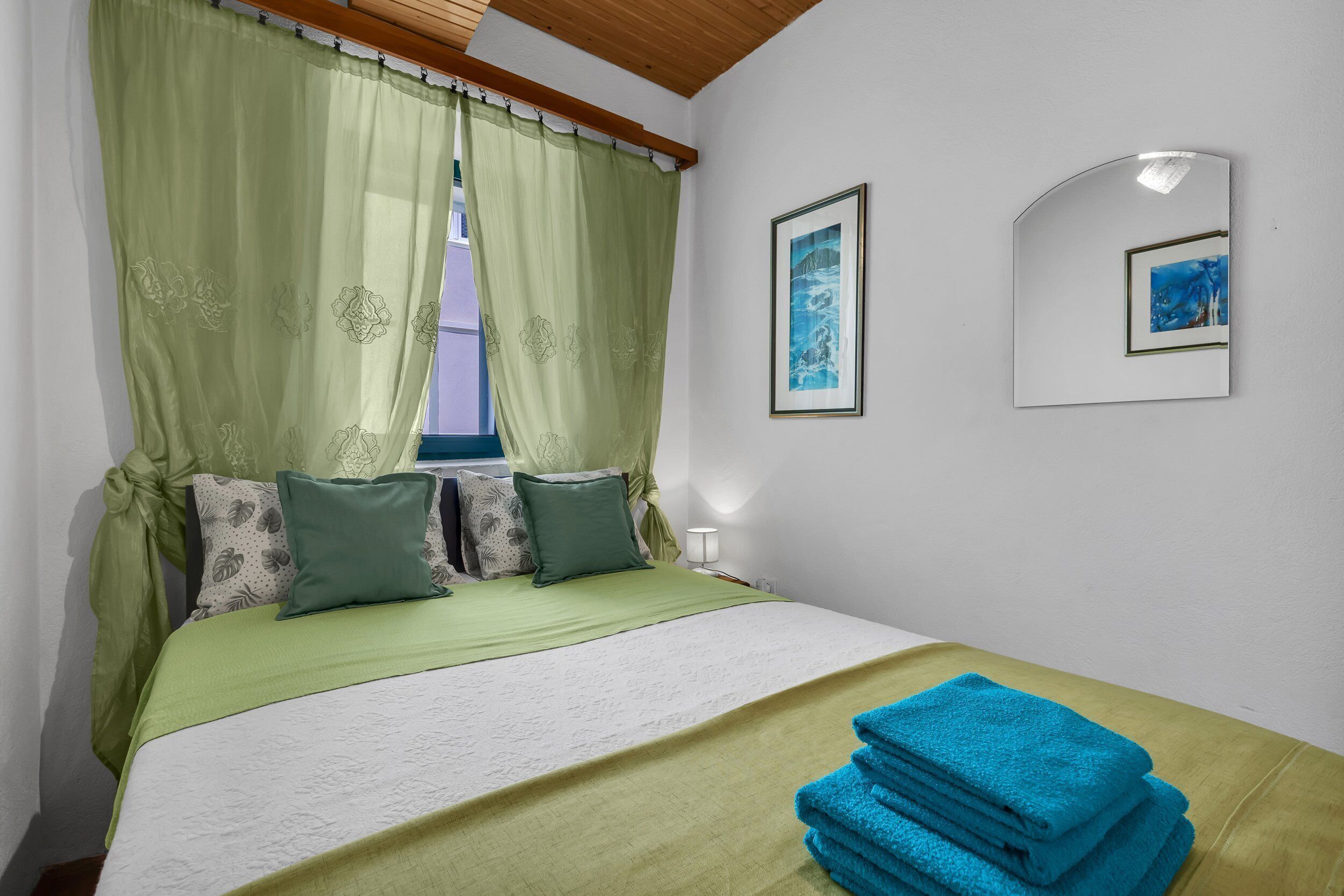 Un lit bien fait, avec une literie verte, une fenêtre ornée de rideaux verts et deux tableaux encadrés accrochés au mur.