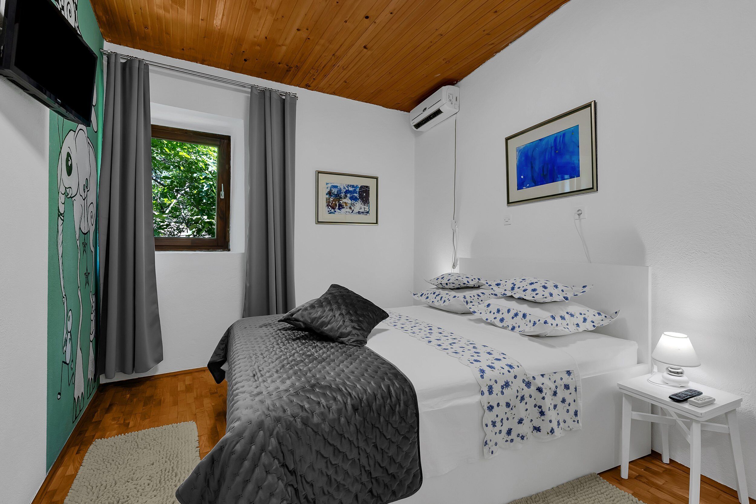 Ein Schlafzimmer mit Bett, Nachttisch mit Lampe, Fenster mit Vorhängen und Bildern an den Wänden.