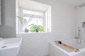 A bathroom with white tiled walls, a window with plants, a bathtub, and a shower.