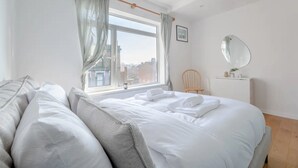 A bedroom with a large bed, a wooden headboard, a window with curtains, a bedside table with a mirror, and a chair. - Peaceful 2 Bedroom Flat With Roof Terrace - Hackney (London)