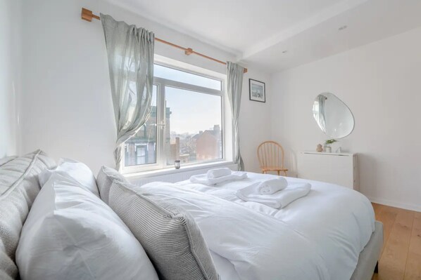 A bedroom with a large bed, a wooden headboard, a window with curtains, a bedside table with a mirror, and a chair.