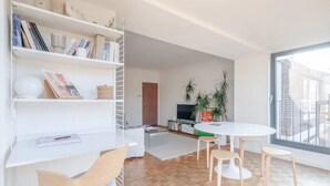 A modern living room with a white bookshelf, a round dining table with four chairs, a TV, and a large window offering a view of the city. - Peaceful 2 Bedroom Flat With Roof Terrace - Hackney (London)