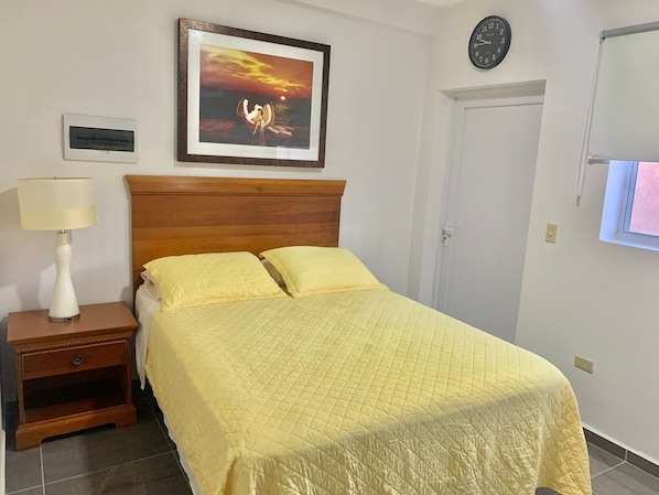 A bedroom with a bed, a nightstand with a lamp, a framed picture on the wall, a clock, and a door.