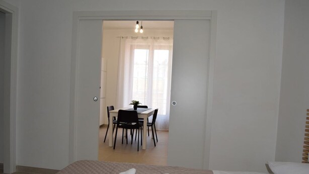 A bedroom with a bed, a bedside table, and a window. There is a door leading to a dining area with a table and chairs. - Brand new apartment and strategically located. (Terrasini)