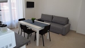 A compact living space with a dining table, four chairs, a grey sofa, a small potted plant, and a flat-screen TV mounted on the wall. - Brand new apartment and strategically located. (Terrasini)