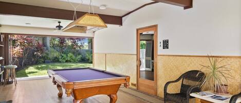 A pool table in a room with wooden flooring, a ceiling fan, and a door leading outside.