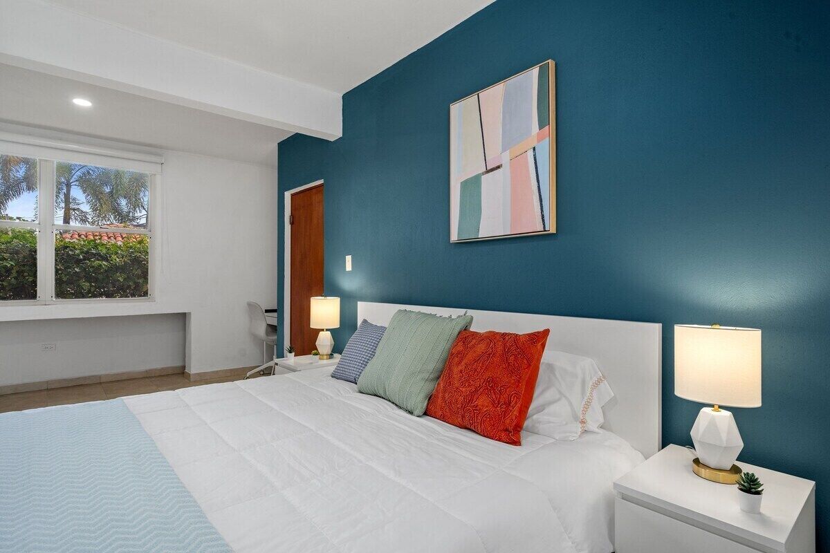 Une chambre avec un grand lit, deux tables de chevet avec des lampes, une fenêtre donnant sur des palmiers et un mur d’accent bleu.