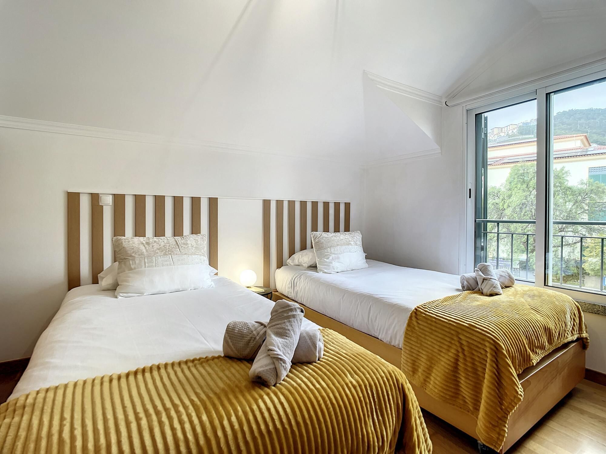 A hotel room with two beds, each with a striped headboard, and a large window offering a view of buildings and trees.