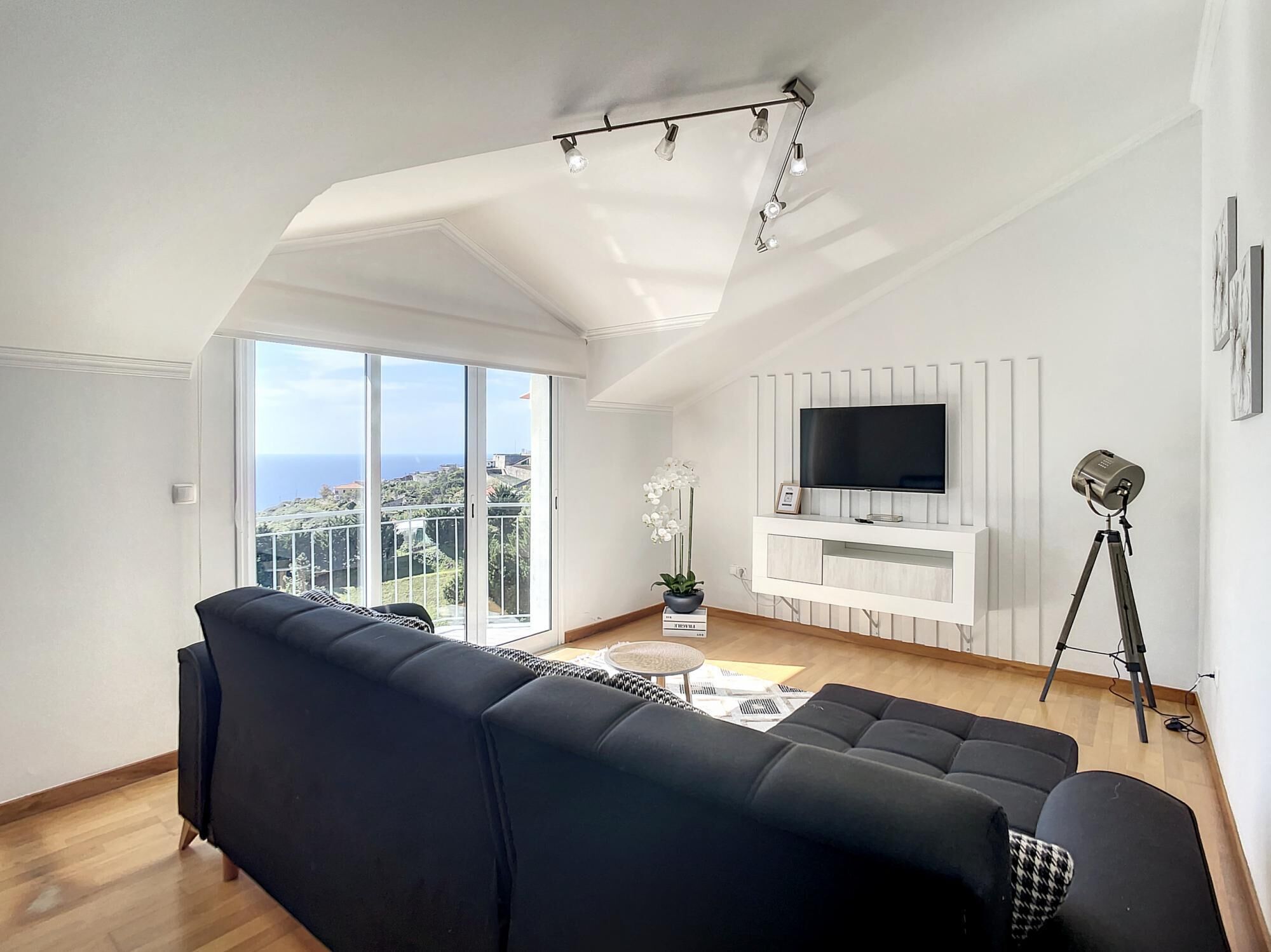A modern living room with a black sofa, a flat-screen TV, a tripod lamp, and a balcony with a view of the sea.