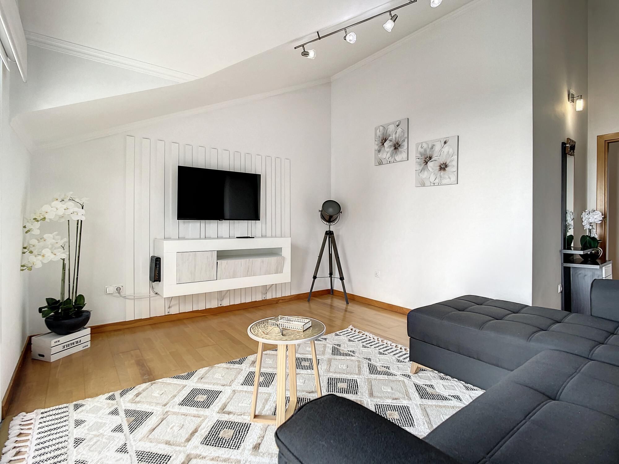Een moderne woonkamer met een grote flatscreen televisie aan een witte muur, een donkergrijze bank, een tapijt met patroon en een kleine ronde tafel.