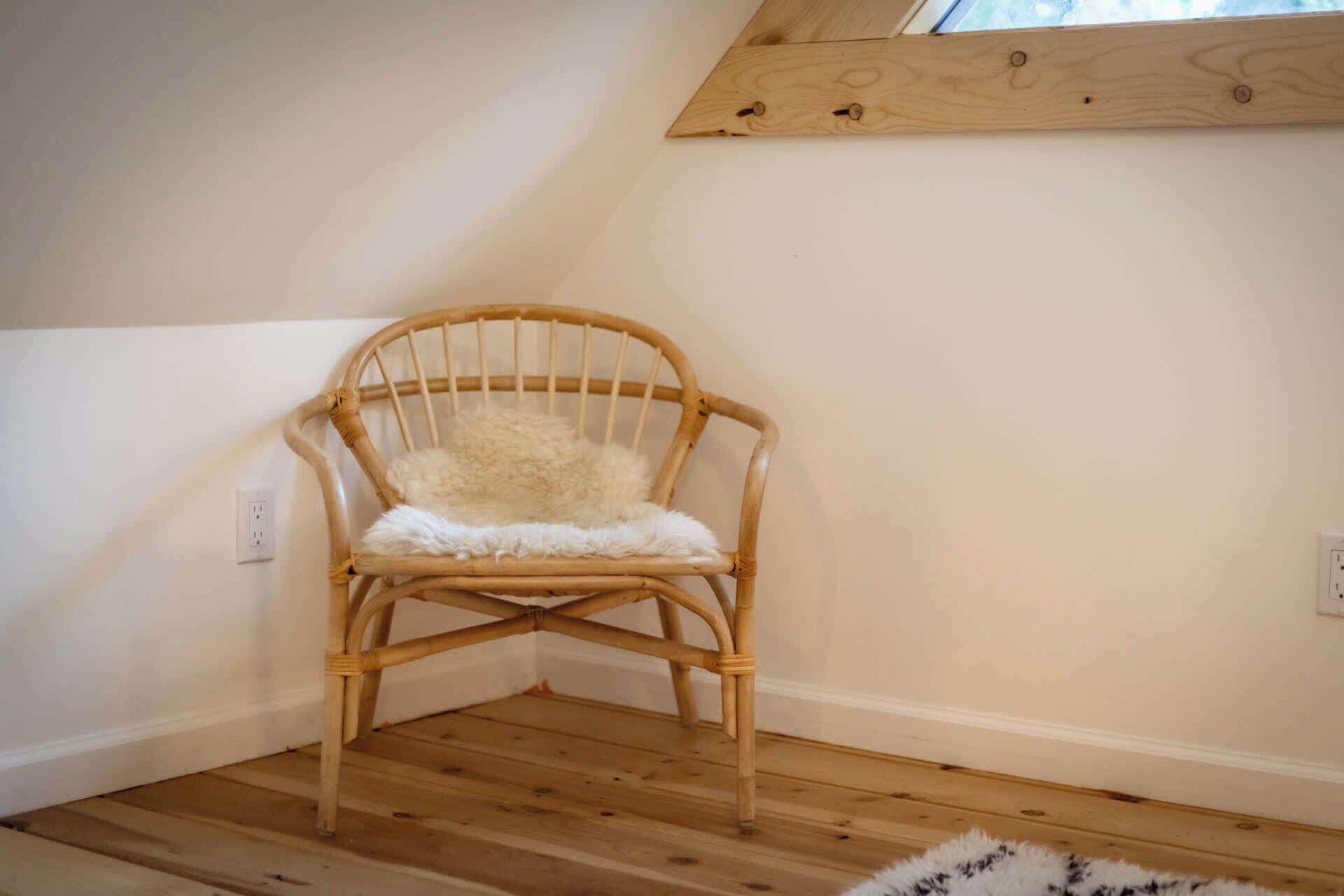 A corner room with a wooden floor, a wicker chair with a fluffy white cushion, and a slanted ceiling.