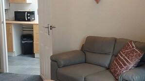 Ein Wohnzimmer mit grauem Sofa, einem pinken Hocker und einer Küchenzeile im Hintergrund.