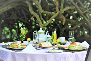 Una mesa dispuesta para una comida, con diversos platos, un juego de té y vasos de bebidas, ubicada al aire libre con árboles de fondo.