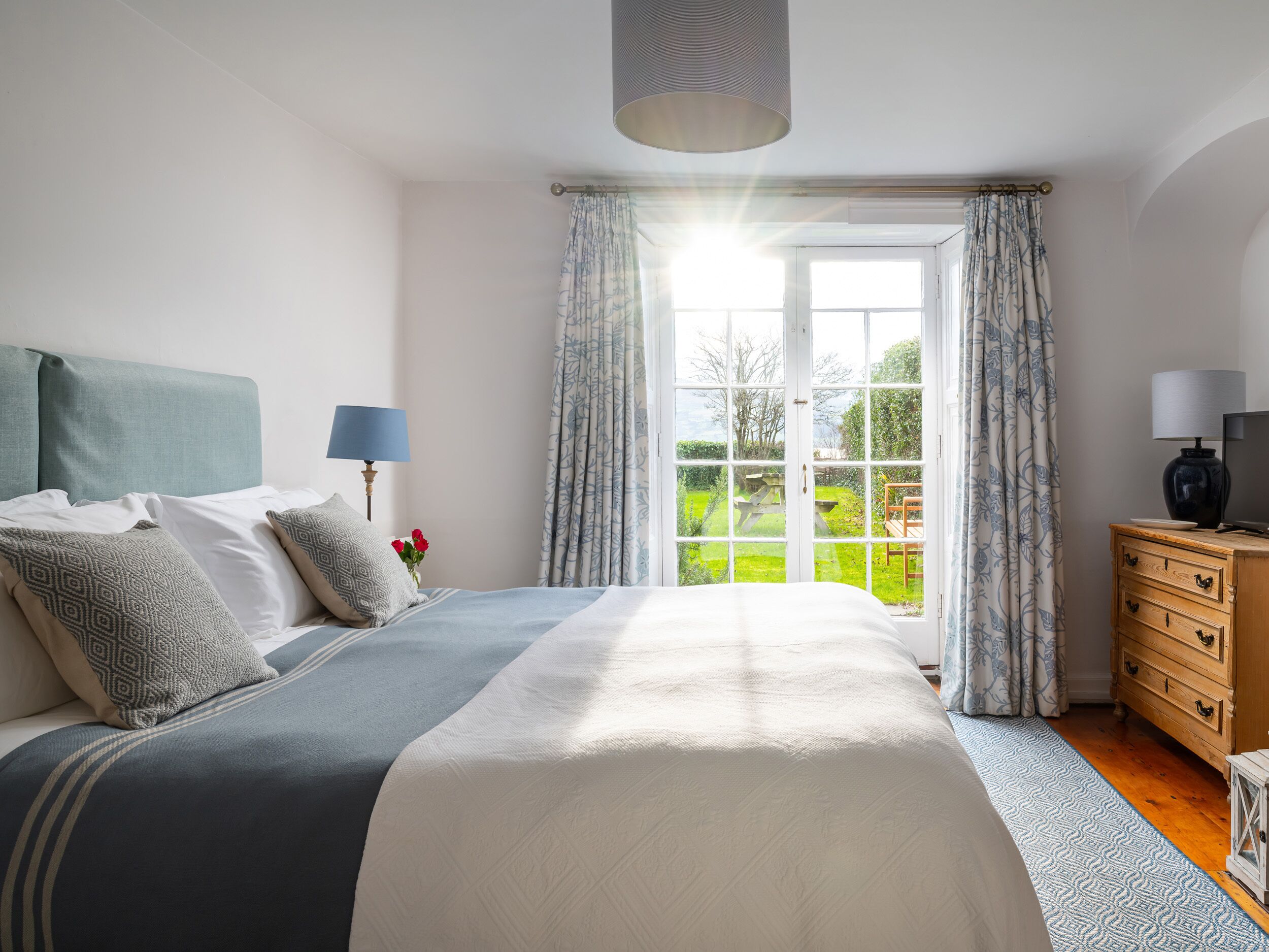 A bedroom with a large bed, a nightstand, a dresser, a lamp, and a view of a garden through a window.