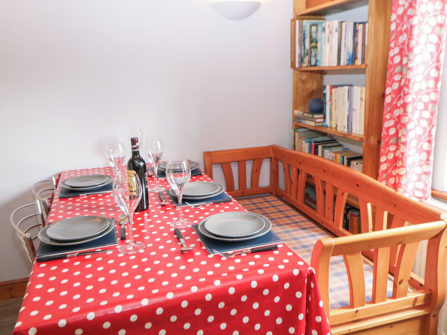 Une table à manger dressée avec des assiettes, des verres et une bouteille, entourée de chaises en bois et, au fond, une bibliothèque.