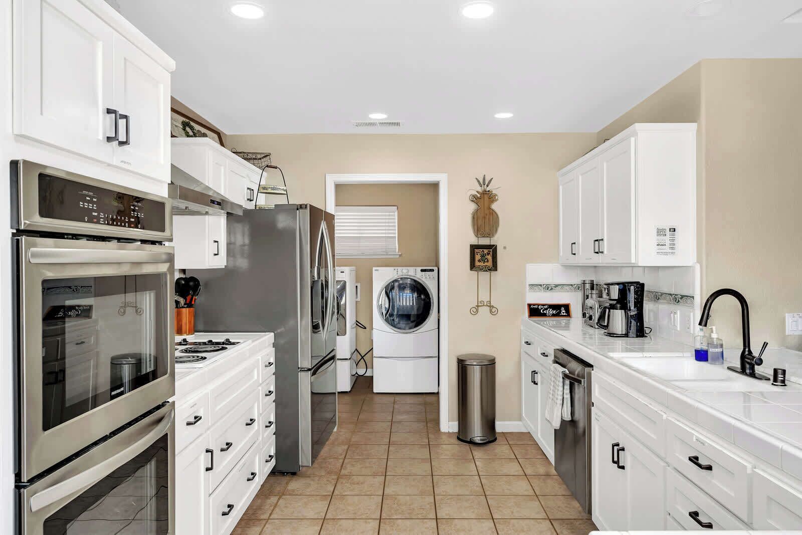 A modern kitchen with white cabinetry, stainless steel appliances, a built-in oven, a refrigerator, a washing machine, and a coffee maker.
