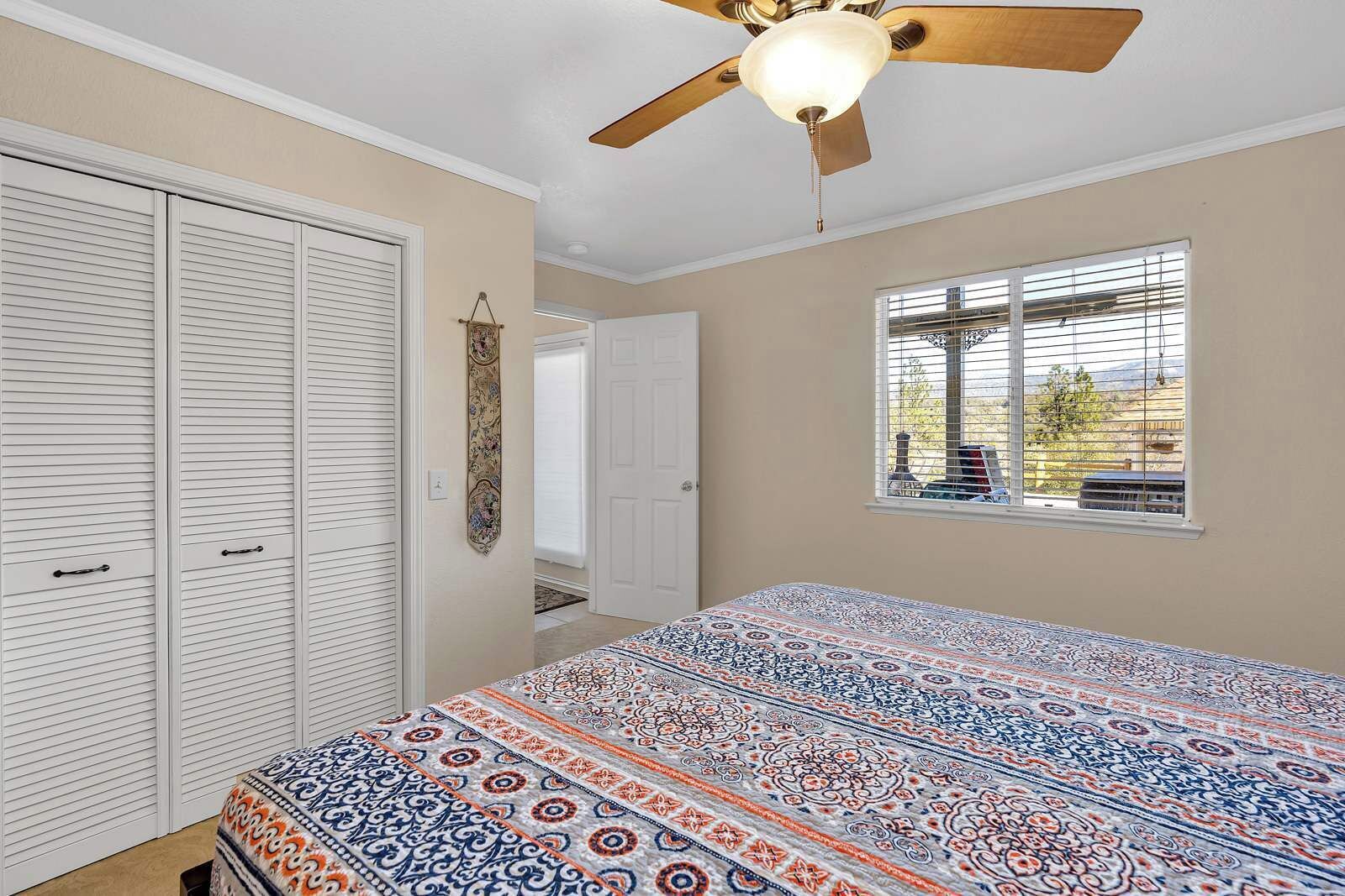 A bedroom with a bed, a ceiling fan, a window with blinds, and a door leading to another room.