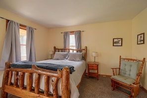 A bedroom with a wooden bed, a wooden chair, a nightstand, and two windows with curtains.