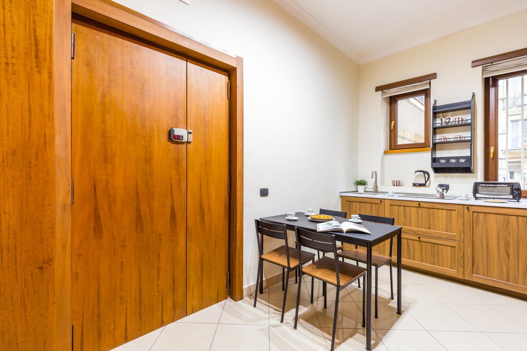 A compact kitchen with wooden cabinets, a dining table with chairs, a window with blinds, and a small countertop with a kettle and a toaster.