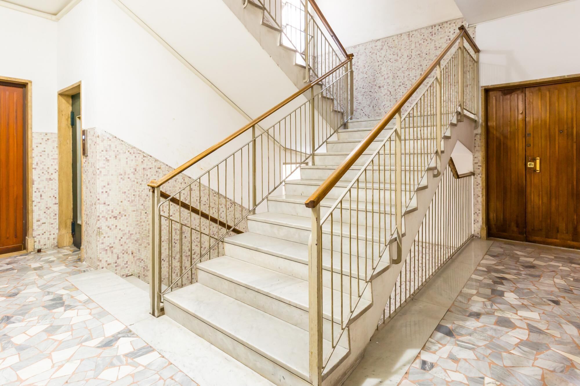 Una escalera con barandillas de metal y pasamanos de madera, que conduce a un suelo de baldosas con un diseño de mosaico.