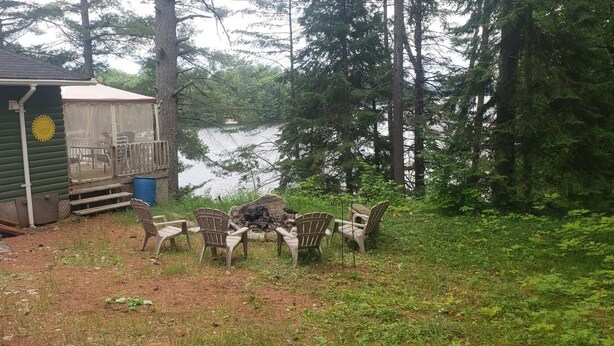 A cabin with a deck overlooking a lake, surrounded by trees. - Quaint Cottage on a Popular Lakefront, Dock, Fire pit. (Kinmount)