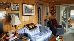 A cozy living room with wooden walls, a sofa, a coffee table, and various items on the table. - Quaint Cottage on a Popular Lakefront, Dock, Fire pit. (Kinmount)