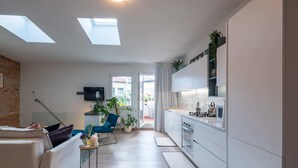 Eine moderne Wohnung mit Dachfenstern, einer Küche, einem Wohnbereich mit einer Couch und einem blauen Sessel, und Blick ins Freie.