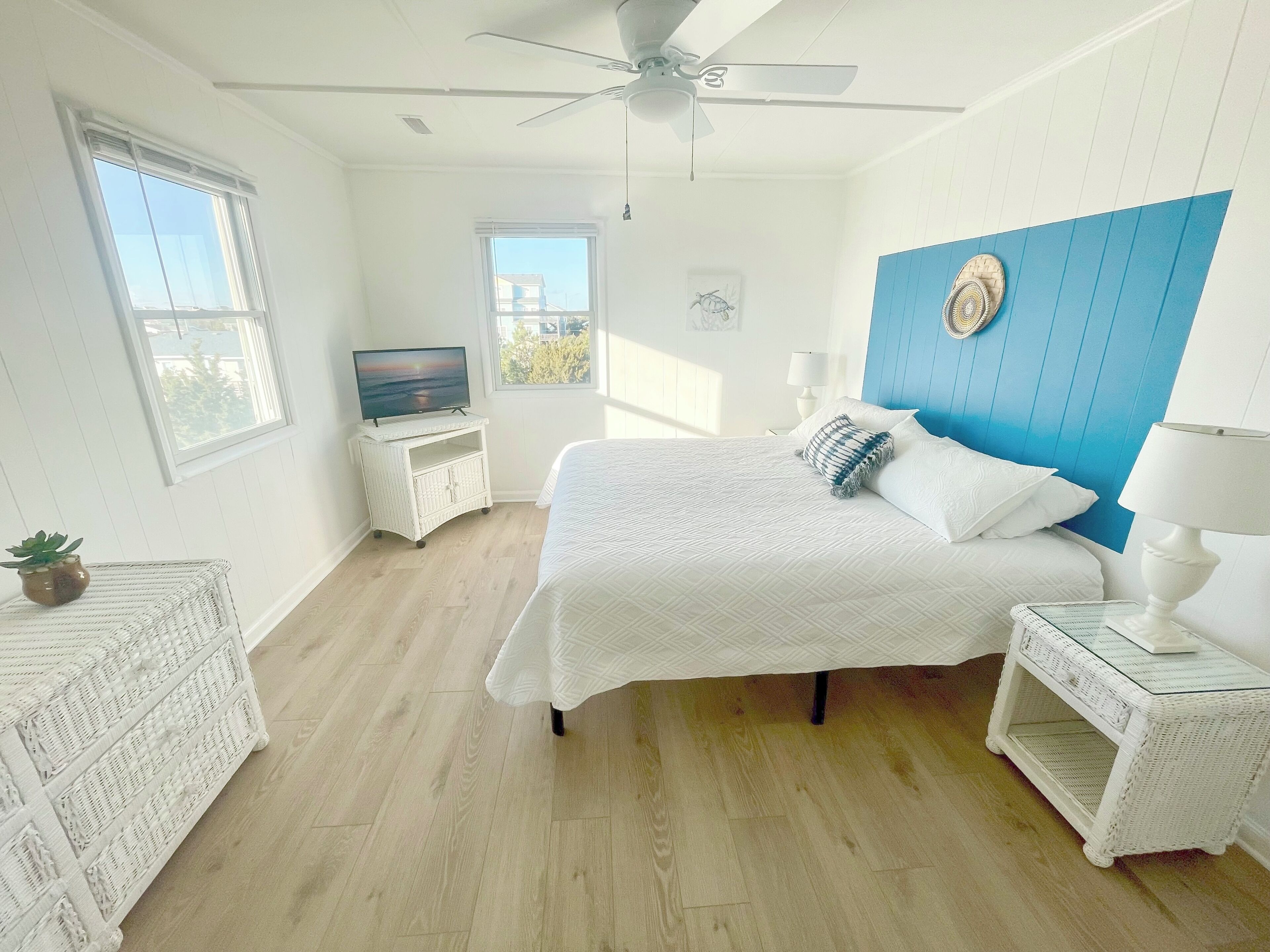 Een slaapkamer met een bed, een televisie op een standaard, een plafondventilator en uitzicht op de omgeving door een raam.