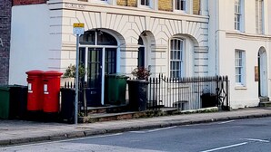 A two-story building with a white facade and a brick section on the left. There are multiple windows, a visible entrance with a door, and a small garden area with a bench. - Captivating 2-bed Apartment in Winchester (Winchester)