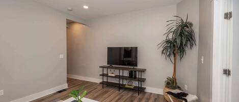 Una sala de estar moderna con un televisor de pantalla plana sobre un mueble negro, piso de madera, pared blanca y una planta grande en maceta.