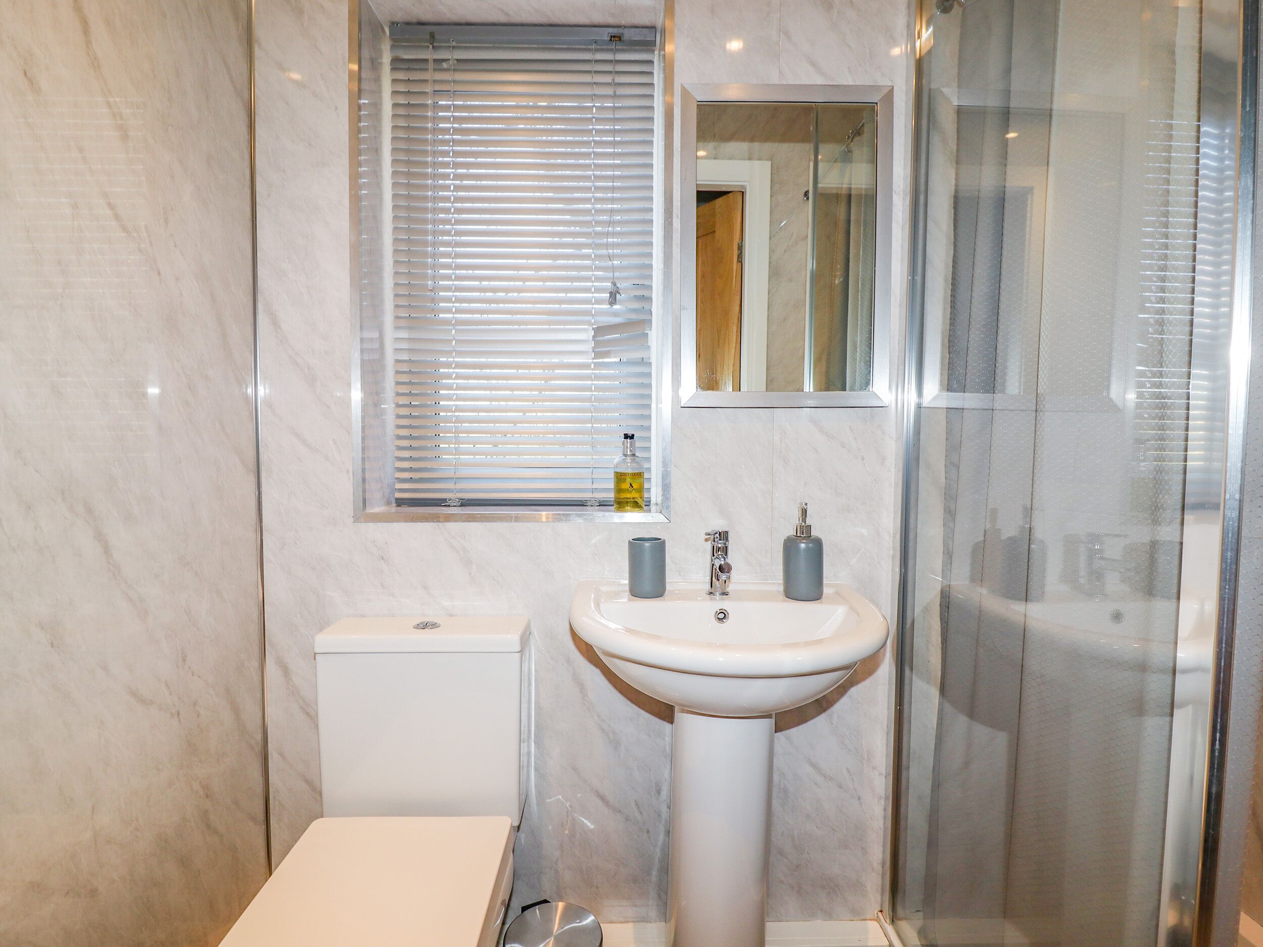 A bathroom with a white pedestal sink, a toilet, a window with blinds, and a glass shower enclosure.
