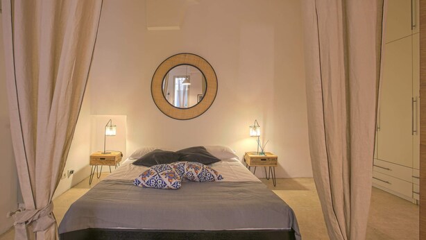 A bedroom with a bed, two bedside tables with lamps, a round mirror on the wall, and a wardrobe. - Santospirito Suite (Florence)