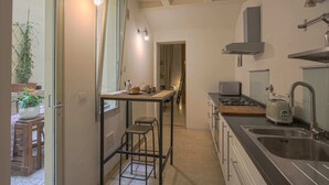 Une cuisine compacte dotée d’un îlot central, d’un évier et d’une cuisinière. On aperçoit un coin repas avec une table et des chaises à travers une porte ouverte.
