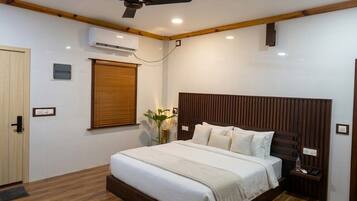Quarto com cama, ventoinha no teto, ar condicionado, janela com persianas e mesa de cabeceira com abajur.