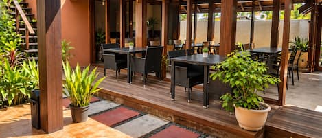 An outdoor patio with wooden tables and chairs, surrounded by potted plants and a tiled floor.