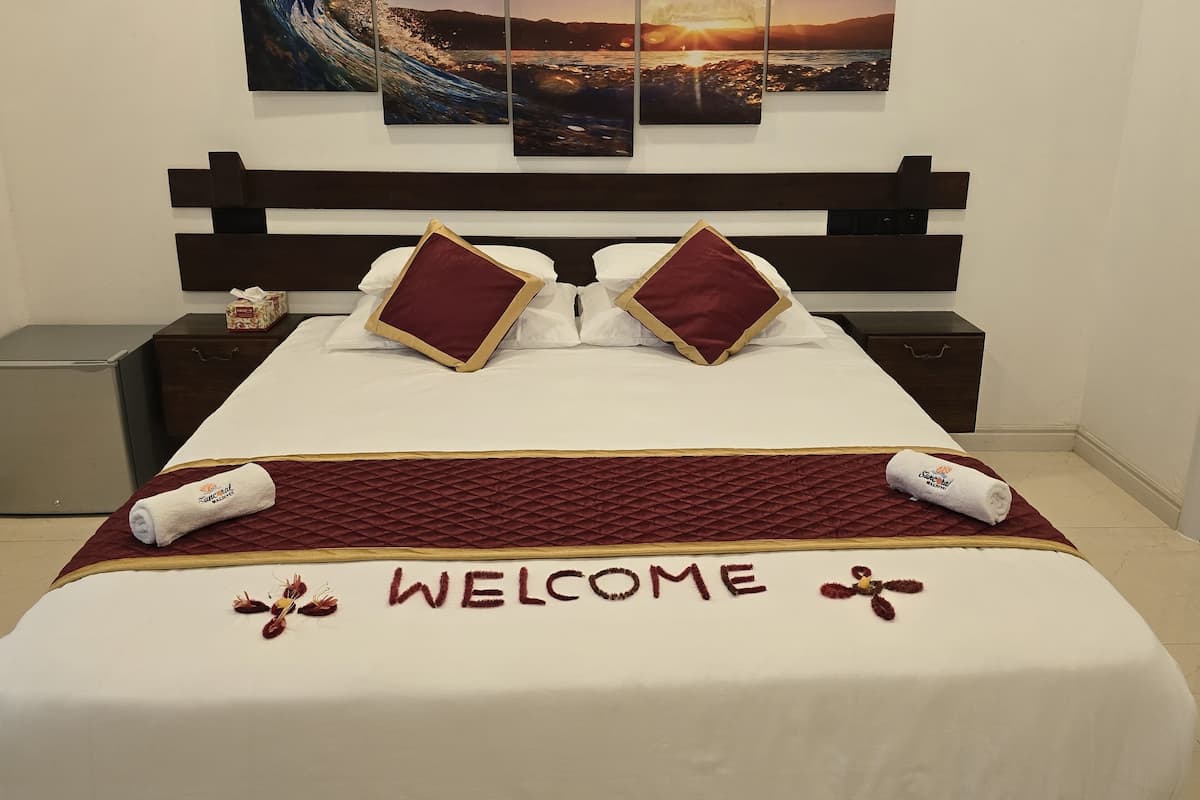 A neatly made bed with a "WELCOME" sign, a nightstand, a small fridge, and a painting of a sunset over water.