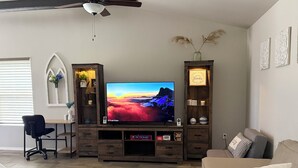 A living room with a flat-screen TV on a wooden entertainment unit, a desk with a chair, and a sofa. - Beautiful and cozy home, perfect for long stay (Alamogordo)