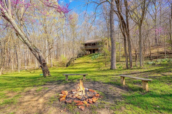 A cabin in a forest with a fire pit and two benches.