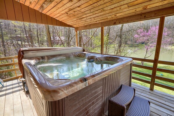A hot tub on a wooden deck with a view of a forest and a pond.